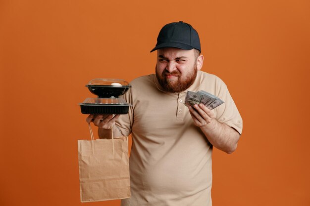 Empleado repartidor con gorra negra y uniforme de camiseta en blanco sosteniendo contenedores de comida con bolsa de papel y dinero en efectivo mirando a un lado insatisfecho haciendo muecas de pie sobre fondo naranja