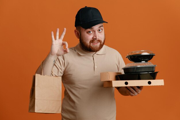 Empleado repartidor con gorra negra y uniforme de camiseta en blanco sosteniendo contenedores de comida con una bolsa de papel haciendo el cartel de ok sonriendo mirando a la cámara de pie sobre fondo naranja