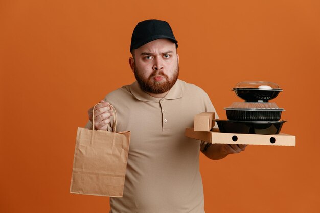Empleado repartidor con gorra negra y uniforme de camiseta en blanco sosteniendo contenedores de comida con una bolsa de papel mirando a la cámara con el ceño fruncido disgustado de pie sobre fondo naranja
