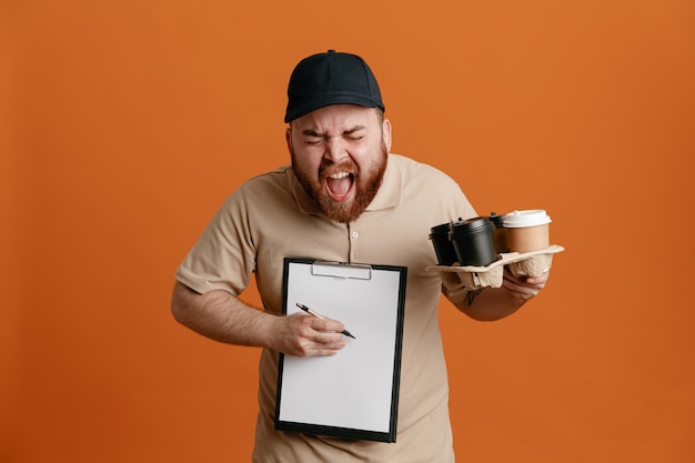 Empleado repartidor con gorra negra y uniforme de camiseta en blanco sosteniendo tazas de café y portapapeles con bolígrafo esperando una firma gritando y gritando enojado y enojado de pie sobre fondo naranja