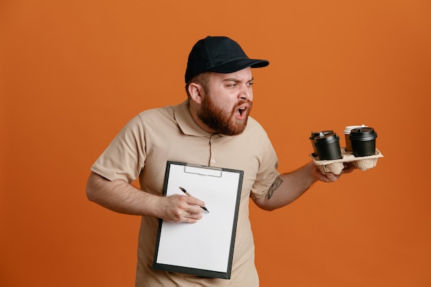 Empleado repartidor con gorra negra y uniforme de camiseta en blanco sosteniendo tazas de café y portapapeles con bolígrafo esperando una firma mirando a un lado confundido y disgustado de pie sobre fondo naranja