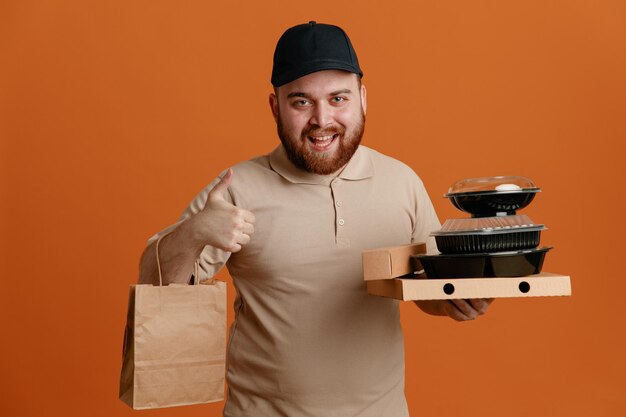 Empleado repartidor con gorra negra y uniforme ts en blanco sosteniendo recipientes de comida con bolsa de papel mirando a la cámara feliz y positivo sonriendo alegremente mostrando el pulgar hacia arriba