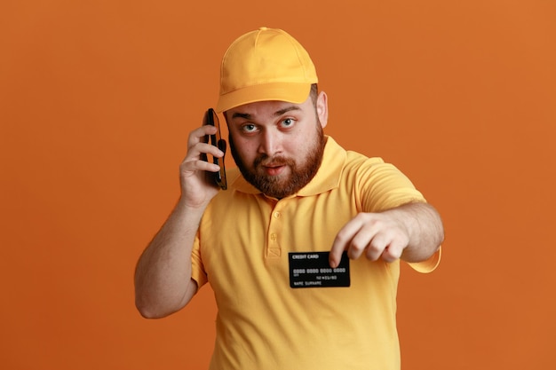 Empleado repartidor con uniforme de camiseta en blanco con gorra amarilla que muestra la tarjeta de crédito hablando por teléfono móvil con aspecto confiado de pie sobre fondo naranja