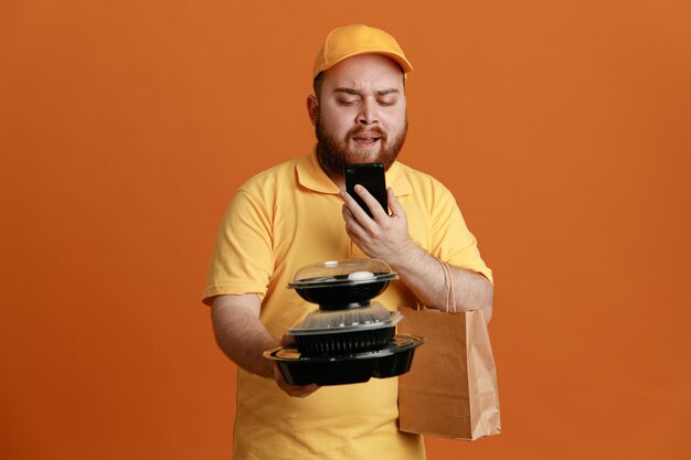 Empleado repartidor en uniforme de camiseta en blanco con gorra amarilla sosteniendo contenedores de comida con una bolsa de papel mirando el teléfono móvil disgustado de pie sobre fondo naranja