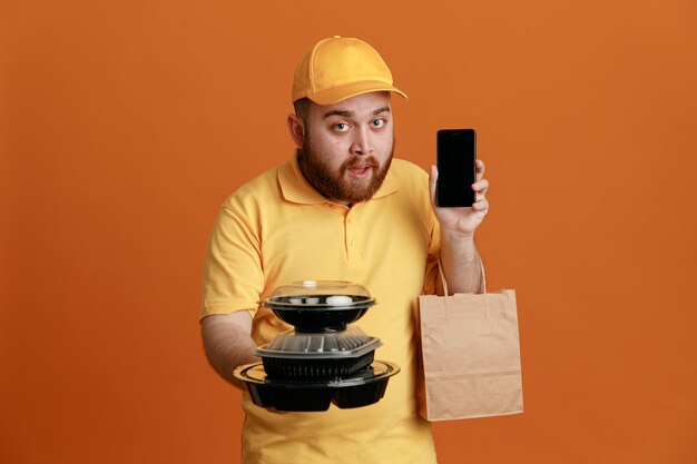 Empleado repartidor en uniforme de camiseta en blanco con gorra amarilla sosteniendo contenedores de comida con una bolsa de papel que muestra el teléfono móvil mirando a la cámara con cara seria de pie sobre fondo naranja