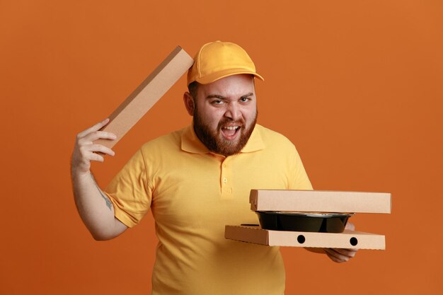 Empleado repartidor en uniforme de camiseta en blanco con gorra amarilla sosteniendo contenedores de comida y caja de pizza mirando a la cámara con expresión molesta de pie sobre fondo naranja