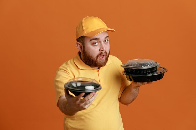 Empleado repartidor en uniforme de camiseta en blanco con gorra amarilla sosteniendo contenedores de comida mirando a la cámara con expresión segura de pie sobre fondo naranja