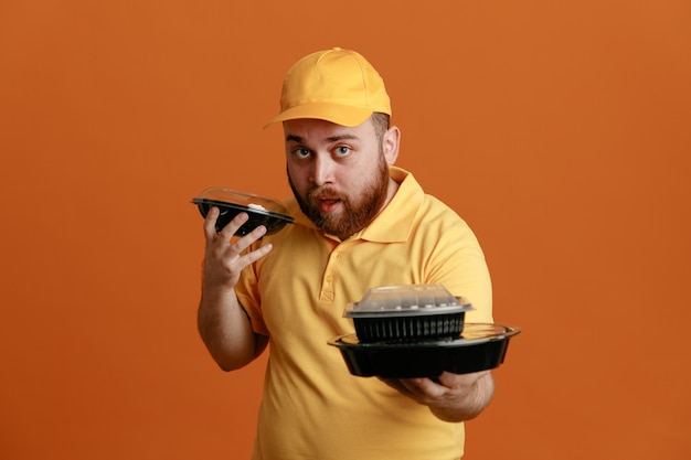 Empleado repartidor en uniforme de camiseta en blanco con gorra amarilla sosteniendo contenedores de comida mirando a la cámara con expresión segura de pie sobre fondo naranja