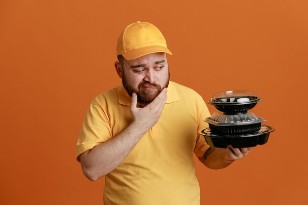 Empleado repartidor en uniforme de camiseta en blanco con gorra amarilla sosteniendo contenedores de comida mirándolos con expresión pensativa pensando de pie sobre fondo naranja