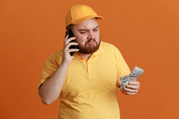 Empleado repartidor en uniforme de camiseta en blanco con gorra amarilla sosteniendo efectivo hablando por teléfono móvil molesto con expresión triste de pie sobre fondo naranja