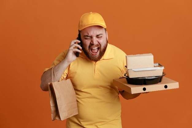 Empleado repartidor con uniforme de camiseta en blanco con gorra amarilla sosteniendo un recipiente de comida y una caja de pizza con una bolsa de papel hablando por teléfono móvil con expresión agresiva de pie sobre un fondo naranja