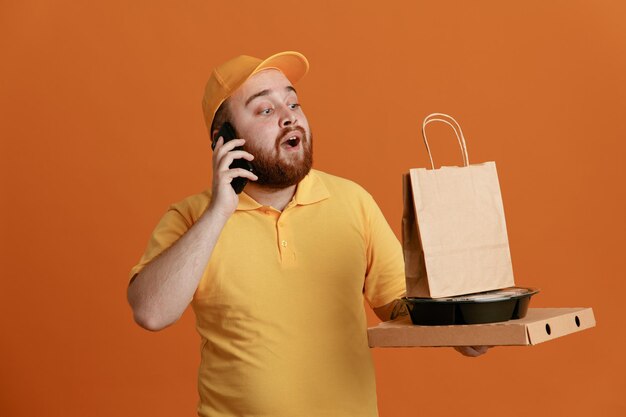 Empleado repartidor en uniforme de camiseta en blanco con gorra amarilla sosteniendo un recipiente de comida y una caja de pizza con una bolsa de papel hablando por teléfono móvil de pie feliz y positivo sobre fondo naranja