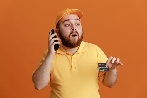 Empleado repartidor en uniforme de camiseta en blanco con gorra amarilla con tarjeta de crédito hablando por teléfono móvil mirando a un lado sorprendido de pie sobre fondo naranja