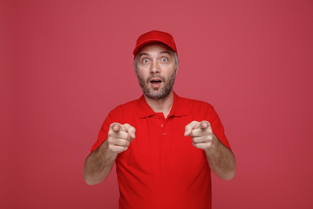 Empleado repartidor con uniforme de camiseta en blanco con gorra roja apuntando con el dedo índice a la cámara feliz y sorprendido de pie sobre fondo rojo