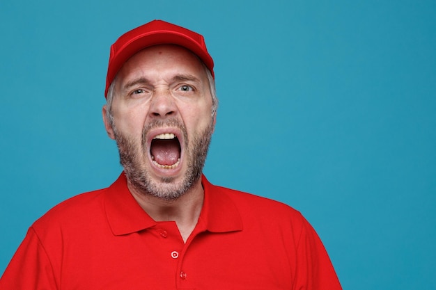 Empleado repartidor con uniforme de camiseta en blanco con gorra roja gritando en voz alta con la boca abierta de pie sobre fondo azul