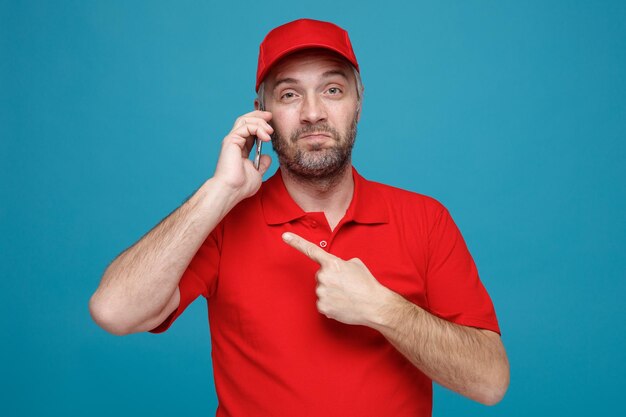 Empleado repartidor en uniforme de camiseta en blanco con gorra roja hablando por teléfono móvil apuntando con el dedo índice al móvil sonriendo feliz y positivo de pie sobre fondo azul