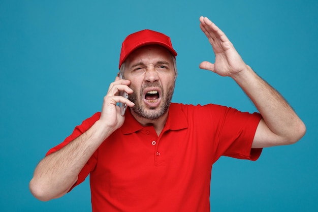 Empleado repartidor en uniforme de camiseta en blanco con gorra roja hablando por teléfono móvil enojado y frustrado levantando el brazo gritando de pie sobre fondo azul