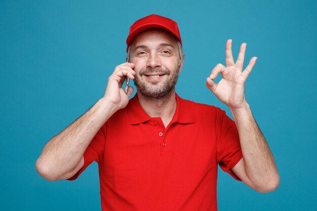 Empleado repartidor en uniforme de camiseta en blanco con gorra roja hablando por teléfono móvil mirando a la cámara sonriendo mostrando el signo ok de pie sobre fondo azul