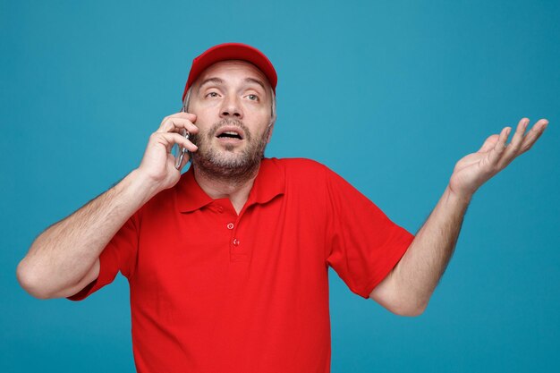 Empleado repartidor en uniforme de camiseta en blanco con gorra roja hablando por teléfono móvil mirando confundido levantando el brazo sin respuesta de pie sobre fondo azul