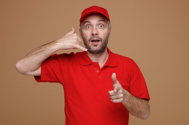 Empleado repartidor con uniforme de camiseta en blanco con gorra roja haciendo gesto de llamarme señalando con el dedo índice a la cámara feliz y sorprendido de pie sobre fondo marrón