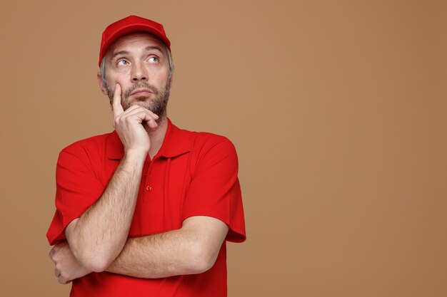 Empleado repartidor en uniforme de camiseta en blanco con gorra roja mirando hacia arriba con expresión pensativa pensando de pie sobre fondo marrón