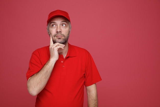 Empleado del repartidor con uniforme de camiseta en blanco con gorra roja mirando hacia arriba pensando desconcertado de pie sobre fondo rojo