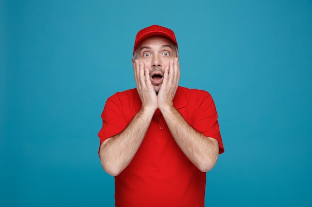 Empleado repartidor en uniforme de camiseta en blanco con gorra roja mirando a la cámara asombrado y sorprendido cubriendo la boca con las manos de pie sobre fondo azul