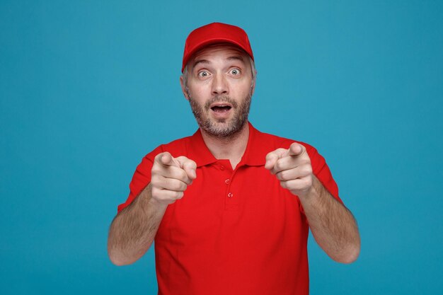 Empleado repartidor con uniforme de camiseta en blanco con gorra roja mirando a la cámara asombrado y sorprendido señalando con los dedos índices con ambas manos a la cámara de pie sobre fondo azul