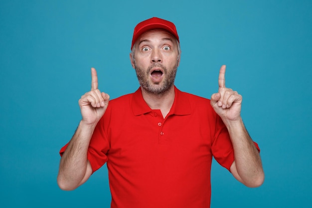 Empleado repartidor con uniforme de camiseta en blanco con gorra roja mirando a la cámara asombrado y sorprendido señalando con los dedos índices hacia arriba con ambas manos de pie sobre fondo azul