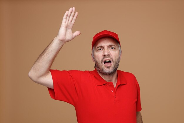 Empleado repartidor en uniforme de camiseta en blanco con gorra roja mirando a la cámara enojado y frustrado gritando levantando el brazo de pie sobre fondo marrón