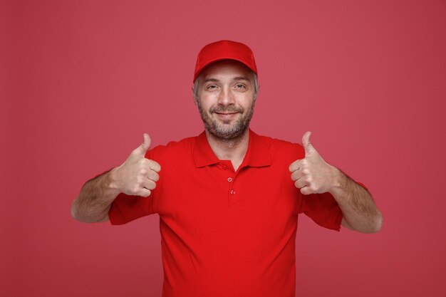 Empleado repartidor en uniforme de camiseta en blanco con gorra roja mirando a la cámara feliz y positivo sonriendo alegremente mostrando los pulgares hacia arriba de pie sobre fondo rojo