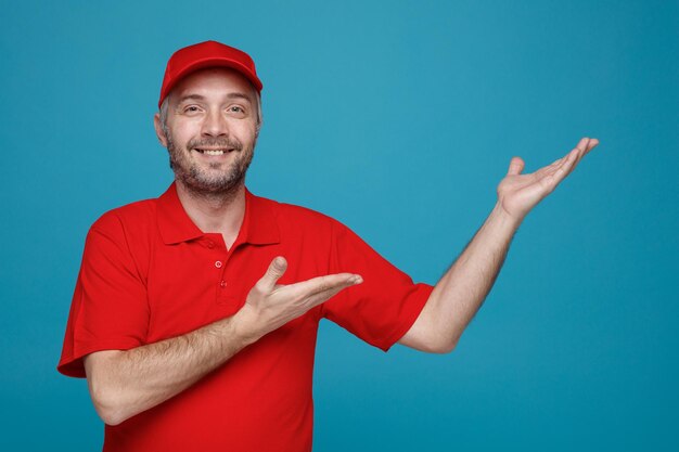 Empleado repartidor en uniforme de camiseta en blanco con gorra roja mirando a la cámara feliz y positivo sonriendo alegremente presentando con los brazos de sus manos algo de pie sobre fondo azul