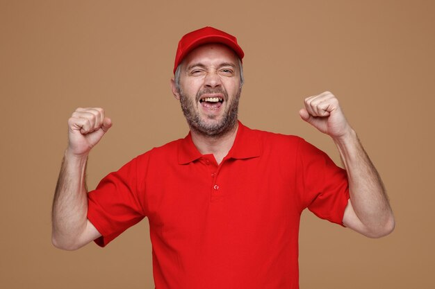 Empleado repartidor con uniforme de camiseta en blanco con gorra roja mirando a la cámara loco feliz y emocionado levantando los puños regocijándose de su éxito de pie sobre fondo marrón