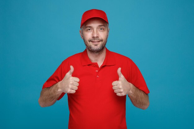 Empleado repartidor en uniforme de camiseta en blanco con gorra roja mirando a la cámara sonriendo feliz y positivamente mostrando los pulgares hacia arriba de pie sobre fondo azul