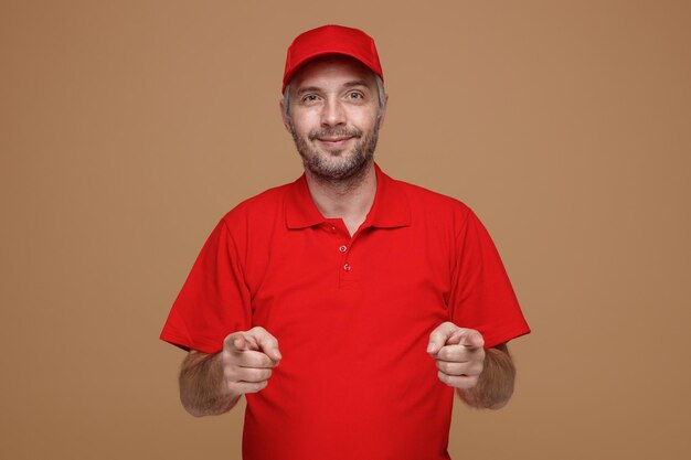 Empleado repartidor con uniforme de camiseta en blanco con gorra roja mirando a la cámara sonriendo feliz y positivamente señalando con los dedos índices con ambas manos a la cámara de pie sobre fondo marrón