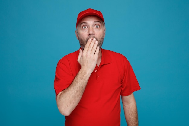 Empleado repartidor con uniforme de camiseta en blanco con gorra roja mirando a la cámara sorprendida cubriendo la boca con la mano de pie sobre fondo azul