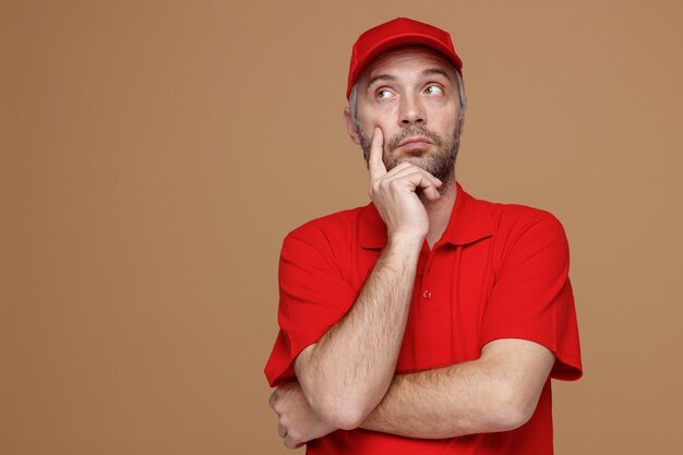 Empleado repartidor con uniforme de camiseta en blanco con gorra roja mirando a un lado con expresión pensativa pensando de pie sobre fondo marrón