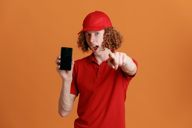 Empleado repartidor con uniforme de camiseta en blanco con gorra roja que muestra el teléfono inteligente apuntando con el dedo índice a la cámara asombrado y sorprendido de pie sobre fondo naranja