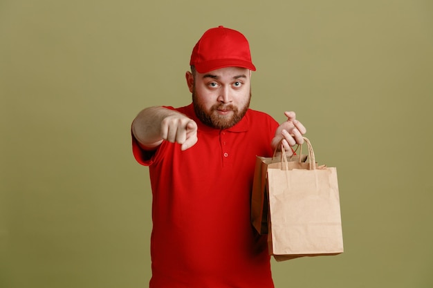 Empleado repartidor en uniforme de camiseta en blanco con gorra roja sosteniendo bolsas de papel apuntando con el dedo índice a la cámara con confianza de pie sobre fondo verde