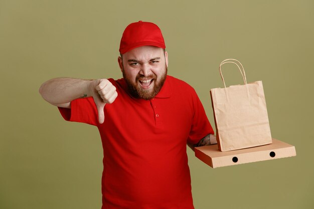 Empleado repartidor con uniforme de camiseta en blanco con gorra roja sosteniendo una caja de pizza y una bolsa de papel mirando a la cámara gritando con expresión molesta mostrando el pulgar hacia abajo de pie sobre fondo verde