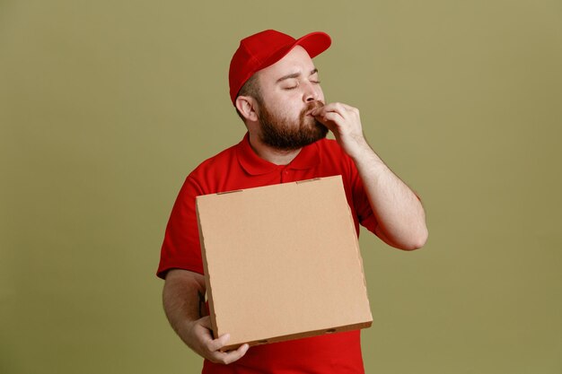 Empleado repartidor en uniforme de camiseta en blanco con gorra roja sosteniendo una caja de pizza que parece feliz y complacido haciendo un gesto excelente de pie sobre fondo verde