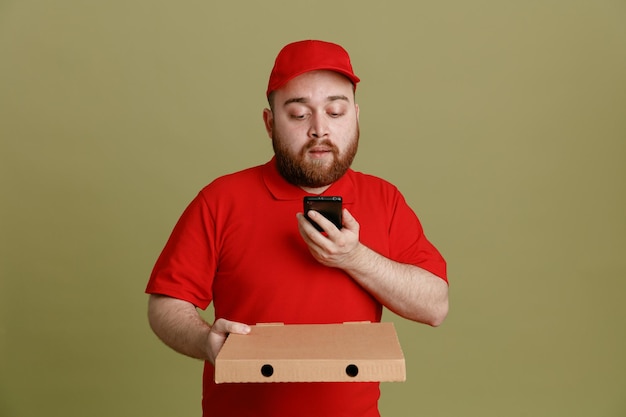 Empleado repartidor en uniforme de camiseta en blanco con gorra roja sosteniendo una caja de pizza y un teléfono inteligente mirando la pantalla desconcertado de pie sobre fondo verde