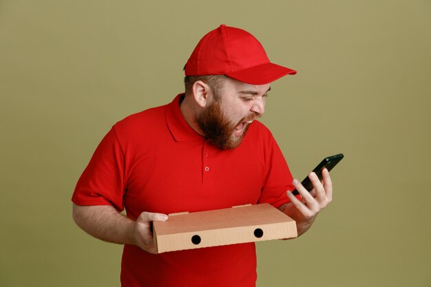 Empleado repartidor en uniforme de camiseta en blanco con gorra roja sosteniendo una caja de pizza y un teléfono inteligente mirando la pantalla gritando con expresión agresiva de pie sobre fondo verde