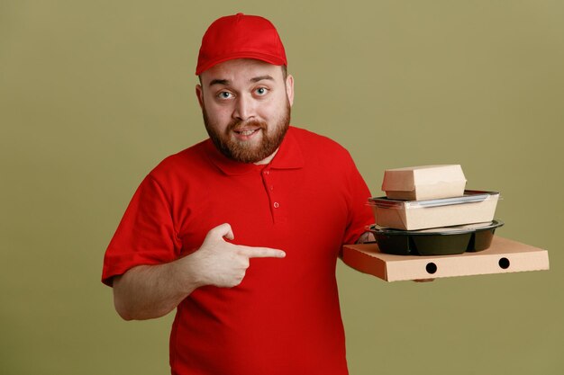 Empleado repartidor con uniforme de camiseta en blanco con gorra roja sosteniendo contenedores de comida y caja de pizza apuntando con el dedo índice sonriendo confiado mirando a la cámara de pie sobre fondo verde