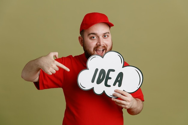 Empleado repartidor con uniforme de camiseta en blanco con gorra roja sosteniendo un discurso de burbuja con una idea de palabra apuntándolo con el dedo índice mirando a la cámara sonriendo alegremente de pie sobre un fondo verde