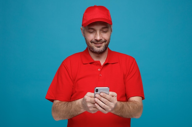 Empleado repartidor en uniforme de camiseta en blanco con gorra roja sosteniendo un mensaje de texto de teléfono inteligente sonriendo alegremente de pie sobre fondo azul