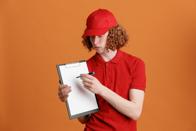 Empleado repartidor en uniforme de camiseta en blanco con gorra roja sosteniendo portapapeles con bolígrafo mirando a la cámara esperando la firma mirando confiado de pie sobre fondo naranja