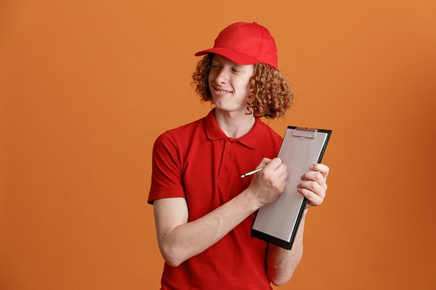 Empleado repartidor en uniforme de camiseta en blanco con gorra roja sosteniendo portapapeles con bolígrafo mirando a un lado esperando la firma sonriendo confiado de pie sobre fondo naranja