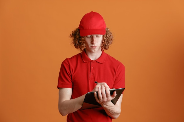 Empleado repartidor en uniforme de camiseta en blanco con gorra roja sosteniendo portapapeles con páginas en blanco haciendo notas que parecen seguros de pie sobre fondo naranja