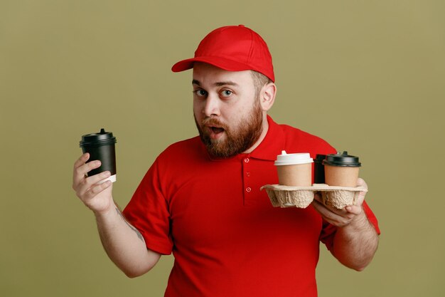 Empleado repartidor en uniforme de camiseta en blanco con gorra roja sosteniendo tazas de café mirando a la cámara asombrado y sorprendido de pie sobre fondo verde